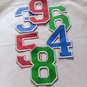 Könnte beinhalten: Eine Sammlung von Stoffaufnähern in Form von Ziffern in Rot, Blau und Grün, jeweils mit einer fetten Blockschrift und einem weißen gestickten Rand. Das Set enthält die Zahlen drei, vier, fünf, sechs, acht und neun in loser Anordnung.
