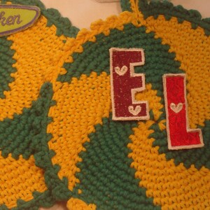 Könnte beinhalten: Zwei gehäkelte Topflappen in Grün und Gelb mit einem Spiralmuster. Die Topflappen haben Filzpatches mit den Buchstaben "E" und "L" darauf. Der Text "Wir sticken" befindet sich auf einem Filzpatch auf einem der Topflappen.