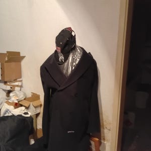 Könnte beinhalten: Ein langer, dunkelblauer, zweireihiger Wollmantel mit breitem Revers und schwarzen Knöpfen, drapiert über einer Form. Eine schwarze Baseballkappe mit rotem und weißem Sticktext liegt auf dem Mantel, der über einer transparenten Kunststoffhülle liegt.