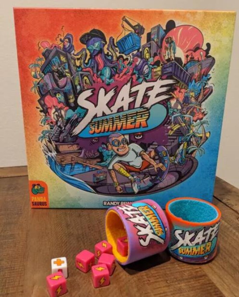 May include: A colorful board game box with the title "Skate Summer" and a cartoon illustration of a skateboarder. The box is on a wooden table with two dice cups and dice. The dice cups are pink and blue with the words "Skate Summer" on them.