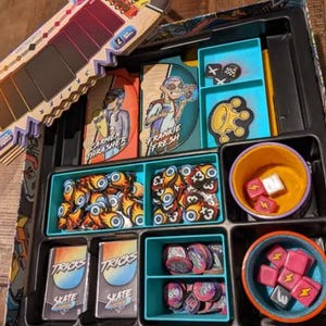 May include: A blue and black game board with various compartments for storing game pieces. The compartments contain small tokens, dice, and cards. The cards have the text "TRICKS" and "SKATE" on them. The board also has two character cards with the text "CARMEN THRASHER" and "FRANKIE FRESH" on them.