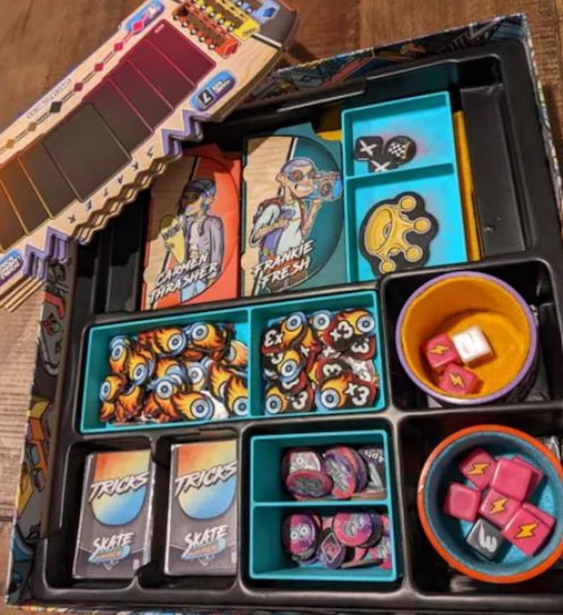 May include: A blue and black game board with various compartments for storing game pieces. The compartments contain small tokens, dice, and cards. The cards have the text "TRICKS" and "SKATE" on them. The board also has two character cards with the text "CARMEN THRASHER" and "FRANKIE FRESH" on them.