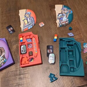 May include: A game with colorful wooden character cards, a red and orange storage tray, and a green and blue storage tray. The character cards feature illustrations of people skateboarding and the text "Carmen Thrasher", "Frankie Fresh", and "Noah Comply".