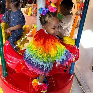 May include: A young girl wearing a rainbow-colored fringed cape and a pink bow in her hair rides a red and yellow carousel horse. The caption "@RoyalPinkBrand" is visible in the image.