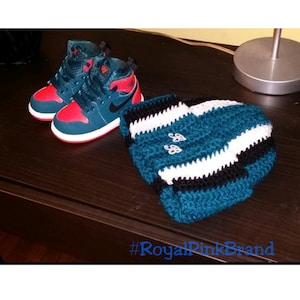 May include: A pair of teal and red baby sneakers with a black swoosh logo, next to a teal, white, and black crocheted hat with the letters "SB" embroidered on it. The text "#RoyalPinkBrand" is visible at the bottom of the image.