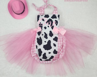 May include: A pink cowboy hat and a romper with a cow print center panel, pink and white gingham sides, and a pink tulle skirt. The romper features the word One in pink script, pink bows at the waist, and tie straps. The text RoyalPinkBrand is visible.