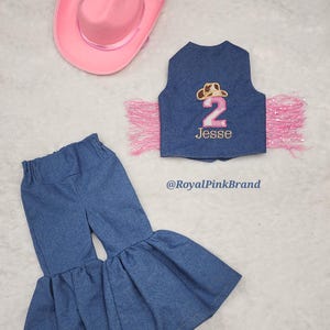 May include: A denim outfit for a child, featuring a vest with pink fringe, a pink cowboy hat, and denim bell bottom trousers. The vest has a pink number 2 and the name "Jesse" embroidered on it. The text "@RoyalPinkBrand" is also visible.