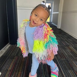 May include: A young child wearing a colorful, fringed jacket, light blue leggings, and matching rainbow-striped slippers. The jacket features a mix of pastel and neon colors. The text "@RoyalPinkBrand" is visible.