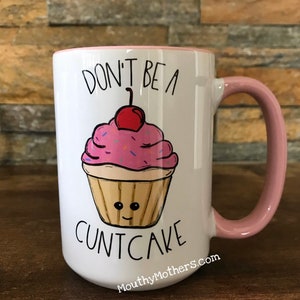 May include: White ceramic coffee mug with a pink rim and handle. The mug has a pink and white cupcake with a cherry on top and the text "Don't be a cuntcake" printed on it. The text "MouthyMothers.com" is printed on the bottom of the mug.