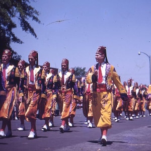 May include: A procession of people in matching yellow and gold outfits with red accents and fezzes. The parade is taking place on a sunny day, with a brick building visible in the background.