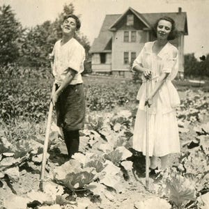 Op de afbeelding: Zwart-witfoto van twee mensen in een tuin. Eén persoon draagt een wit overhemd en een donkere broek en houdt een tuingereedschap vast. De andere persoon draagt een witte jurk. Een huis staat op de achtergrond.
