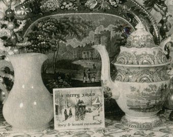 May include: A still life photograph featuring a vintage teapot, a pitcher, and a Christmas card with the text "a merry xmas". The teapot and pitcher are light-colored, and the teapot has a detailed pattern. The card reads "mary & leonard via northwood".