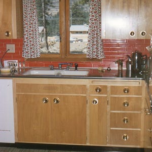 May include: A vintage kitchen with light wood cabinets and drawers, featuring brass hardware. Red tile backsplash and a window with floral curtains. The kitchen includes a sink, countertop, and a retro-style floor.