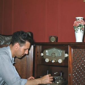 Peut inclure: Une console radio en bois vintage dotée d'un cadran central avec trois boutons de commande, d'une grille de haut-parleur à motif losange et d'une surface supérieure portant un vase blanc avec des détails de roses roses et une petite horloge carrée.