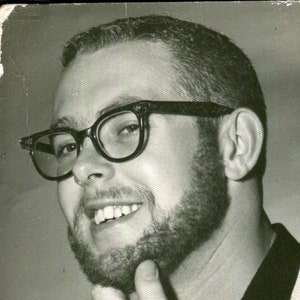 May include: Black and white photo of a man with a beard and glasses. He is smiling and has his hand on his chin.