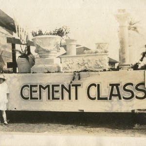 May include: Vintage black and white photograph of a parade float. The float features cement sculptures, including planters and a column. The words "CEMENT CLASS" are painted on the front. A child in a hat stands in front of the float.