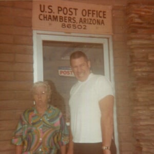 May include: Vintage photograph of two people standing in front of a U.S. Post Office in Chambers, Arizona. The sign above the door reads "U.S. Post Office Chambers, Arizona 86502". The woman is wearing a colorful patterned blouse.