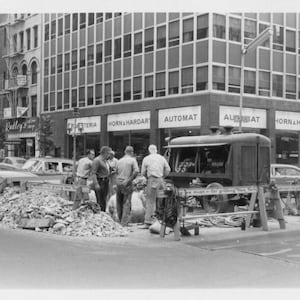 May include: Black and white photograph of a street scene from July 1959. A group of men are gathered around a piece of machinery on a city street. Buildings and cars are visible in the background.
