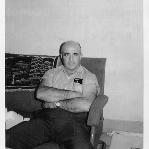 Könnte beinhalten: Ein Schwarzweißfoto eines Mannes, der auf einem Stuhl sitzt und die Arme verschränkt. Er trägt ein helles Hemd und eine dunkle Hose. Der Stuhl hat einen gemusterten Stoffbezug.