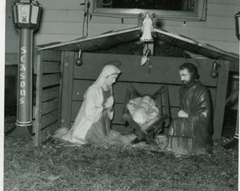 May include: Black and white photograph of a Christmas nativity scene. The scene includes figures of Mary, Joseph, and baby Jesus in a manger. A lamppost with the word "Seasons" is visible. The year "1956" is written at the bottom.