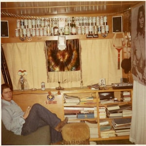 May include: A vintage interior scene with a man seated on a couch. The room features a shelf filled with books, a tie-dye tapestry, and a collection of beer cans. A poster of a woman is on the wall. The scene evokes a retro, possibly 1970s, aesthetic.