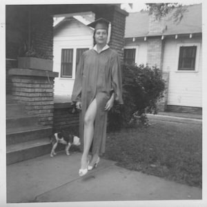 May include: Black and white photograph of a person in a graduation gown and cap, standing on a concrete walkway. A small dog is visible near the steps of a brick house. The person is wearing high heels.