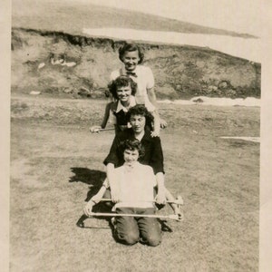 May include: Vintage black and white photograph of four people posing outdoors. The photo is dated 1950 and features handwritten text at the top. The people are arranged in a line, with the bottom person kneeling and holding a wooden frame.