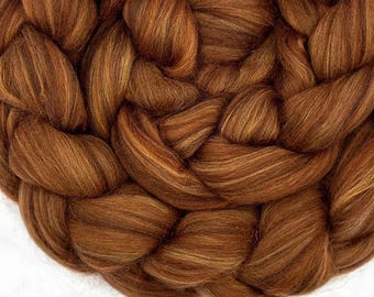 May include: A thick, chunky braid of soft, unspun wool roving in a rich, warm auburn brown colour. The fibres are densely packed and intertwined in a circular pattern, showcasing a smooth, fibrous texture with subtle variations in copper tones.
