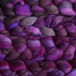 Könnte beinhalten: Ein dickes, grob gestricktes Textil mit einem sich wiederholenden Zopfmuster. Das Material zeigt eine abwechslungsreiche Farbpalette aus tiefem Violett, Magenta und gedämpften bräunlich-grauen Tönen, die eine weiche, strukturierte Oberfläche bilden.