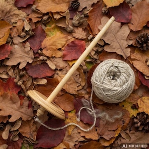 May include: A light-coloured wooden drop spindle with a smooth, tapered shaft and a circular whorl, positioned next to a round ball of thick, textured, off-white yarn that has a loose strand trailing from its centre.