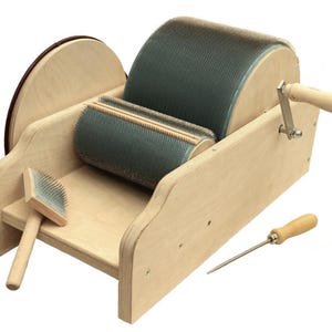 Könnte beinhalten: Eine Wollkardiermaschine aus Holz mit einer großen rotierenden Trommel und einer kleineren Walze, beide mit feinen Metallzähnen bedeckt. Eine Handkurbel und zwei Handkarden sind enthalten. Die Maschine ist aus hellem Holz gefertigt.