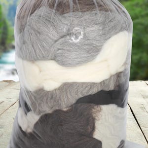 Könnte beinhalten: Ein klarer Plastikbeutel gefüllt mit Wollvlies in verschiedenen Farbtönen. Die Farben umfassen Weiß, Hellgrau, Mittelgrau, Dunkelgrau und Braun. Der Beutel ist oben mit einem roten Bindfaden verschlossen. Im Hintergrund ein verschwommenes Bild von Fluss und Bäumen.