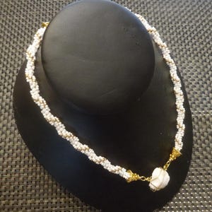 May include: A white and gold beaded necklace with a white pendant. The necklace is on a black display stand.