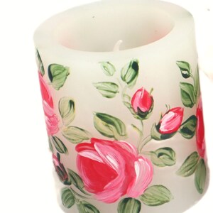 May include: A white pillar candle with hand-painted red roses and green leaves. The floral design wraps around the candle, creating a decorative accent. The candle has a single wick.