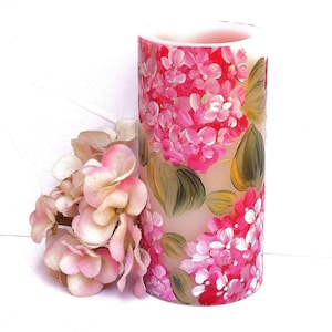 Peut inclure: Une bougie peinte à la main avec un motif floral. La bougie est blanche avec des fleurs roses et vertes peintes dessus. La bougie est cylindrique et a un diamètre d'environ 7,5 cm.