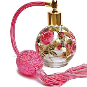May include: A pink glass perfume bottle with a gold top and a pink tassel. The bottle is decorated with hand-painted pink roses and green leaves. The bottle has a gold chain with a pearl charm hanging from it.