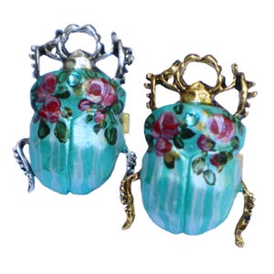 May include: Two beetle-shaped brooches featuring a bright turquoise body with white vertical stripes and a floral pattern of pink roses and green leaves. One brooch has a silver-toned metal frame and legs, while the other has a gold-toned metal frame.