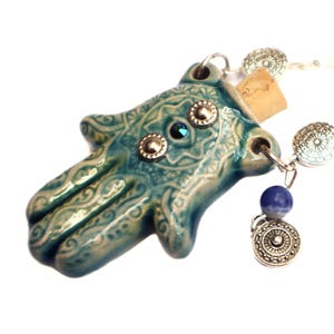 May include: A turquoise ceramic Hamsa hand pendant with silver accents and a blue gemstone. The pendant is attached to a silver chain with decorative beads and a small blue and silver charm. The Hamsa hand has an intricate design.