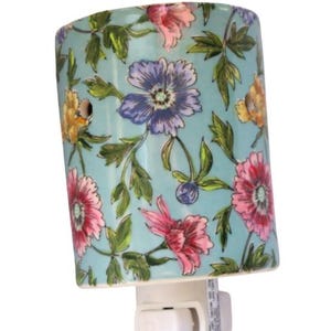 Puede incluir: Una luz de noche de cerámica con un diseño floral. La luz tiene un fondo azul claro con flores rosas, moradas y amarillas y hojas verdes. La luz de noche se enchufa directamente a una toma de corriente y tiene un interruptor de encendido/apagado.