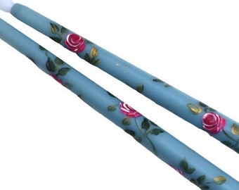 May include: Two blue candles with white wicks and a floral design of pink roses and green leaves.