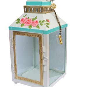 Puede incluir: Linterna de metal blanco con un borde azul turquesa y un diseño floral. La linterna tiene una ventana de vidrio y una cadena dorada con un borla de cuentas doradas.