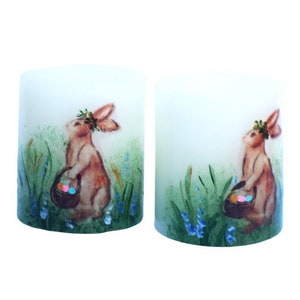 May include: Two white pillar candles featuring a brown bunny rabbit holding a basket of colourful Easter eggs. The bunnies are surrounded by green grass and blue flowers.