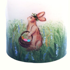 May include: A hand-painted candle featuring a brown bunny holding a basket of colourful Easter eggs. The bunny is wearing a flower crown and is surrounded by green grass and blue flowers.