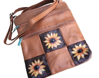 May include: A brown leather crossbody bag with three zippered compartments and a patchwork design featuring painted sunflowers.