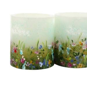 Könnte beinhalten: Zwei zylindrische Kerzen mit handgemaltem Blumendesign. Die Kerzen haben einen hellgrünen Hintergrund mit einer Wiese aus bunten Blumen und grünem Gras, die um den Sockel gemalt sind. Die Kerzen sind unbeleuchtet.