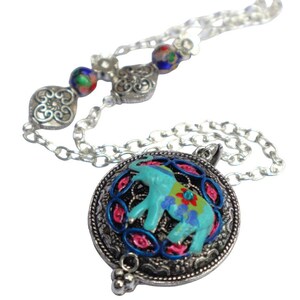 May include: A silver chain necklace with a round pendant featuring a blue elephant with colorful details. The pendant is surrounded by a silver filigree design. The necklace also has two silver beads with blue and white enamel.