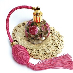 May include: A pink glass perfume bottle with a gold top and a pink tassel. The bottle is decorated with painted roses and a pearl. The bottle is sitting on a crocheted doily.