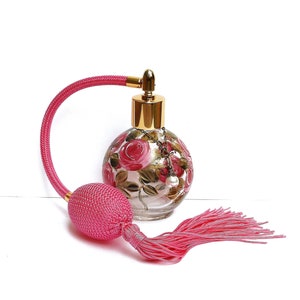 May include: A pink glass perfume bottle with a gold top and a pink tassel. The bottle is decorated with hand-painted pink roses and green leaves.