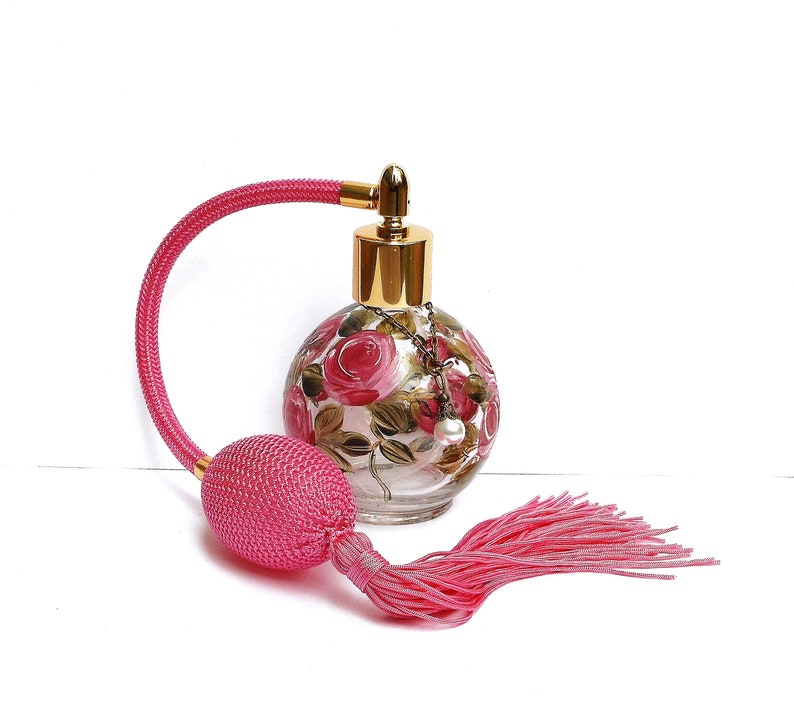 May include: A pink glass perfume bottle with a gold top and a pink tassel. The bottle is decorated with hand-painted pink roses and green leaves.