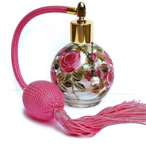 May include: A pink glass perfume bottle with a gold cap and a pink tassel. The bottle is decorated with hand-painted pink roses and green leaves. The bottle has a gold chain with a pearl charm.